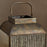 A close-up of a vintage-style metal lantern with a distressed bronze finish, featuring a curved top handle and ribbed front panels, set against a solid brown background.